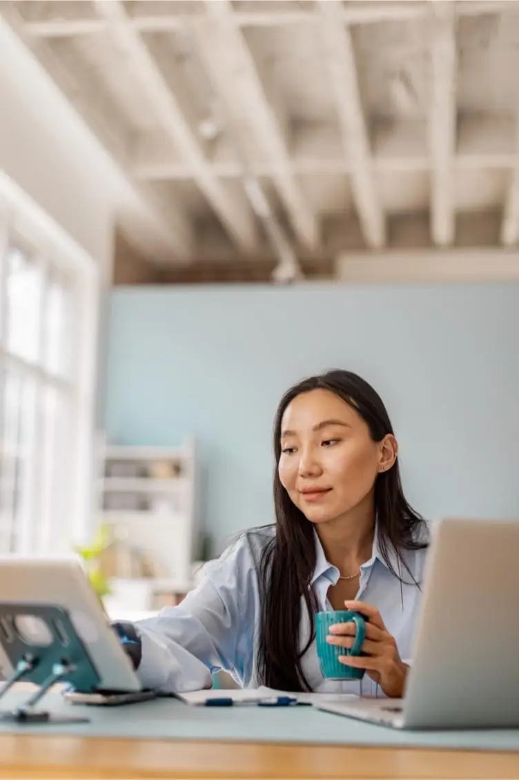 A woman drinking from a cup while working on a tablet in a bright room.